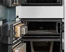 MARELI SYSTEM / CALDAIA LEGNA LCG LAMBDA - L’immagine illustra il piano inferiore del vano di carico dove è posta la feritoia di passaggio della fiamma e si intravede la piastra superiore di posizionamento. Sui lati destro e sinistro si notano i convogliatori dell’aria comburente secondaria e, prestando attenzione, si vedono i vari fori che la distribuiscono uniformemente sui due lati. Sulla sinistra si vede anche lo sportello aperto su ci è posizionato il dispositivo dell’accensione automatica